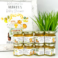 Honey jars with personalized labels for a baby shower, next to a decorative sign and plant.
