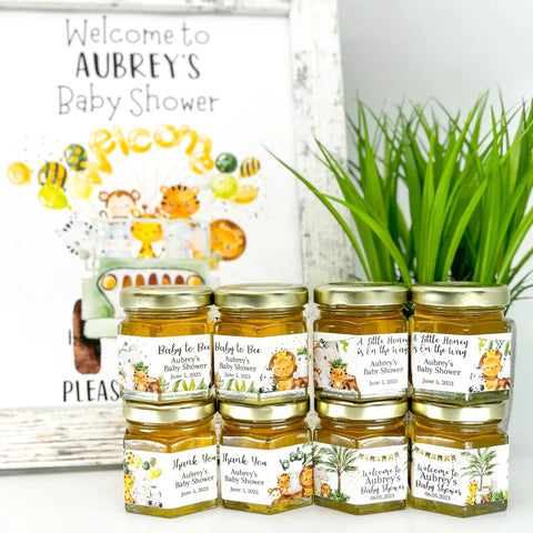 Honey jars with personalized labels for a baby shower, next to a decorative sign and plant.