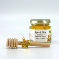 Jar of honey with a wooden dipper and gold flower charm on a white background