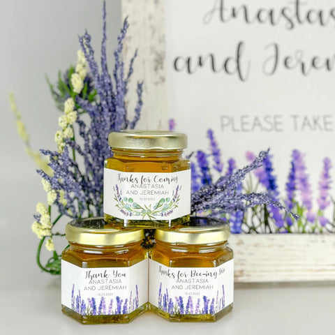 Three jars of honey with labels on a white surface with lavender flowers in the background.