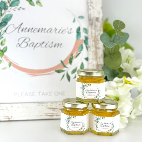 Three small jars of honey with personalized labels on a white surface, with a decorative sign in the background.