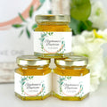 Three jars of honey with personalized labels on a white surface with flowers in the background