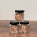Three hexagonal glass jars with black lids containing a pinkish salt on a wooden surface.