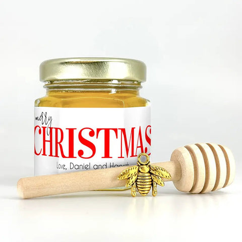 Jar of honey with a gold lid and 'Merry Christmas' label, accompanied by a wooden honey dipper with a gold bee charm on a white background.