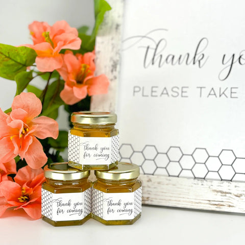 Three jars of honey with 'thank you for coming' labels next to pink flowers and a sign.
