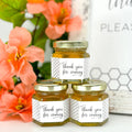 Three small hexagonal jars of honey with 'thank you for coming' labels, surrounded by pink flowers.