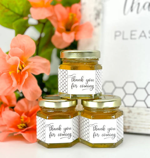 Three small hexagonal jars of honey with 'thank you for coming' labels, surrounded by pink flowers.