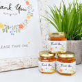 Three jars of honey with 'Thank You' labels on a white surface with a plant in the background.