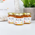 Three jars of honey with 'Thank You' labels on a white surface, featuring a plant and decorative elements in the background.