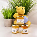Yellow teddy bear with honey jars on a white surface with plants in the background