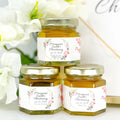 Three small jars of honey with personalized labels on a white surface with flowers in the background