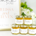 Small jars of honey with personalized labels on a white surface with flowers in the background.