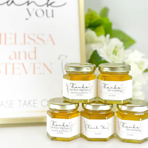 Small jars of honey with personalized labels on a white surface with flowers in the background.