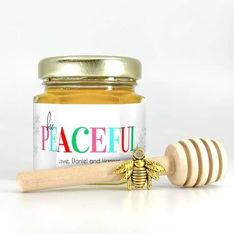 Jar of honey with a colorful label, wooden honey dipper, and gold bee charm on a white background