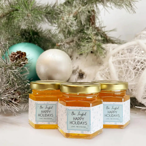 Three jars of honey with 'Bee Joyful' labels in front of Christmas decorations.