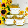 Three jars of honey with personalized labels surrounded by sunflowers on a white surface.
