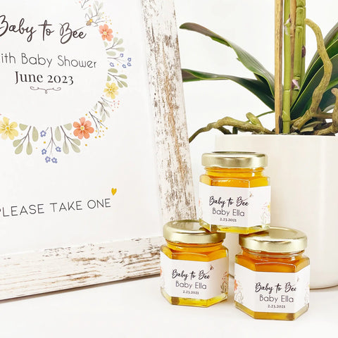 Jars of honey with personalized labels on a white surface with a plant in the background.