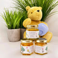 Three jars of honey with personalized labels, a teddy bear, and a plant in the background.