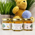 Three small jars of honey with personalized labels in front of a teddy bear holding a blue jar of honey