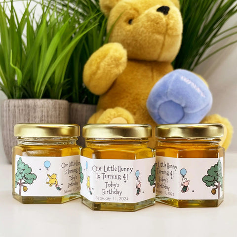 Three small jars of honey with personalized labels in front of a teddy bear holding a blue jar of honey