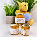 Three jars of honey with baby shower labels and a teddy bear holding a blue gift bag in front of plants.