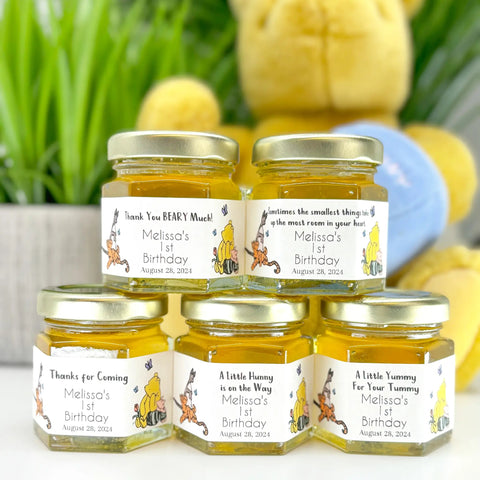 Five small jars of honey with personalized labels on a white surface, with a teddy bear and greenery in the background.