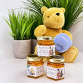 Three jars of honey with personalized labels next to a teddy bear and plants on a white surface