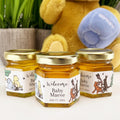 Three small jars of honey with personalized labels in front of a plush toy and plant.