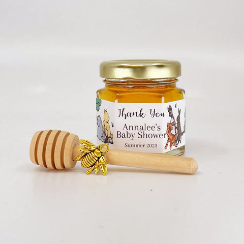 Jar of honey with a gold lid, gold bee charm, and wooden honey dipper on a white background