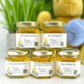 Five jars of honey with personalized labels on a white surface, with a blurred background.