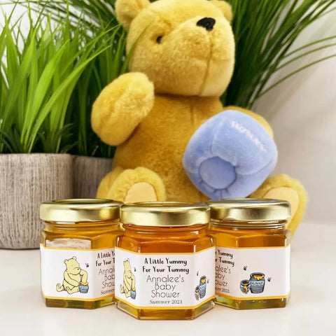 Three jars of honey with personalized labels in front of a teddy bear holding a baby blanket.