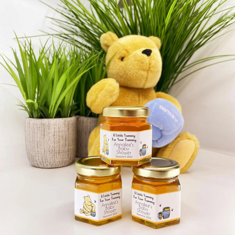 Three jars of honey with labels and a teddy bear holding a blue honey pot in front of plants.