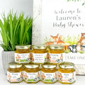 Jars of honey with personalized labels for a baby shower, placed on a white surface with a plant in the background.