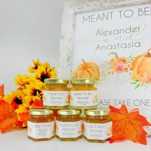 Jars of honey with personalized labels in front of a decorative sign with pumpkins and flowers.