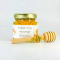 Jar of honey with a gold lid, 'thank you' label, gold bee charm, and wooden honey dipper on a white background