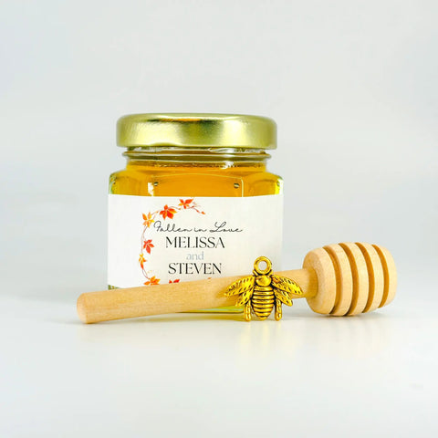 Jar of honey with a gold lid, gold bee charm, and wooden dipper on a white background