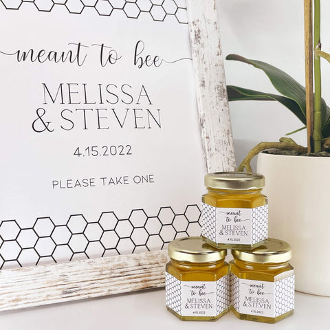 Honey jars with personalized labels next to a sign with 'meant to bee' text, on a white surface with a plant in the background.