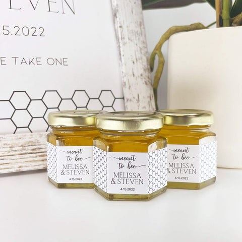 Three jars of honey with personalized labels on a white surface and sign in background