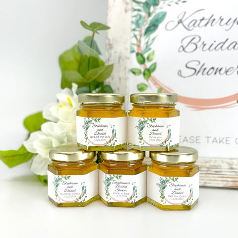 Jars of honey with personalized labels on a white surface with flowers and a sign in the background.