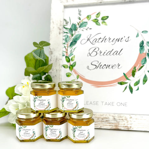 Jars of honey with personalized labels with a bridal shower sign and flowers in the background
