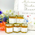 Small jars of honey with personalized labels, honey, and gold lids. Title options include Bee Our Guest, Found My Honey, Bride to Bee, Welcome, Love is Sweet, and more. Showcasing different flower color options: multi, yellow, blue, red/pink, and purple.