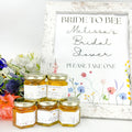 Small jars of honey with personalized labels and gold lids in front of bridal shower sign. Label titles include Bee Our Guest, Bride to Bee, Found My Honey, Welcome, Love is Sweet, and more. Floral color options shown: red/pink, purple, yellow, blue, and multi. 
