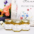 Three jars of honey with 'Meant to Bee' labels on a white surface, surrounded by flowers.