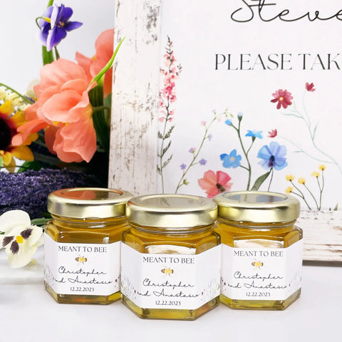 Three jars of honey with 'Meant to Bee' labels on a white surface, surrounded by flowers.