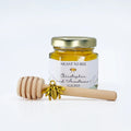 Jar of honey with a gold lid, gold bee charm, and wooden honey dipper on a white background