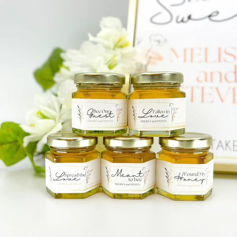Five jars of honey with labels on a white surface with flowers in the background
