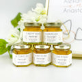 Five jars of honey with personalized labels on a white surface with flowers in the background.