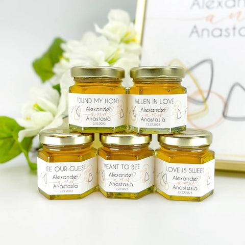 Five jars of honey with personalized labels on a white surface with flowers in the background.