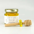 Jar of honey with a gold lid, gold 'love' charm, and wooden dipper on a white background