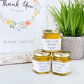Three jars of honey with labels on a white surface with a plant in the background.